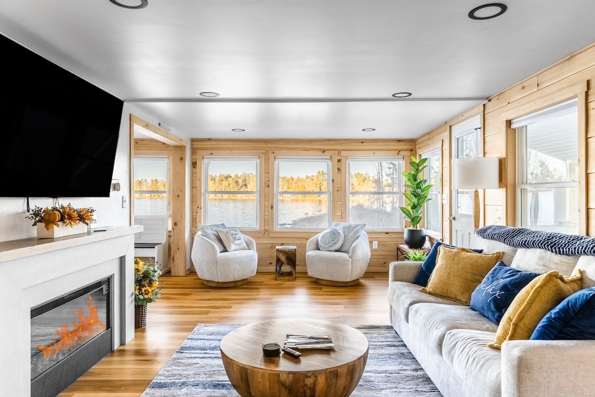 An open-concept living area features a cozy sofa with an array of cushions, complemented by two plush armchairs. A circular coffee table is positioned at the center, while a fireplace adds warmth. Large windows frame scenic waterfront views, inviting natural light into the space.