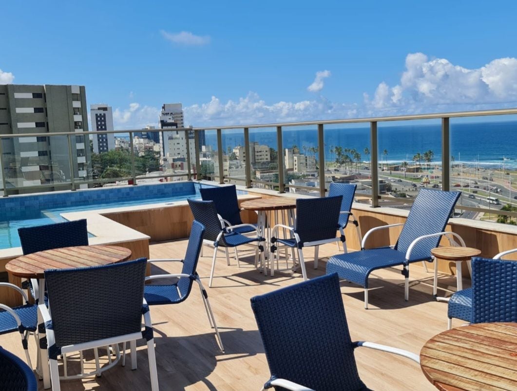 The rooftop area features a selection of modern, blue chairs and wooden tables, arranged for relaxation and socializing. A swimming pool can be seen in the background, with a view of the ocean and cityscape beyond, under a bright blue sky.