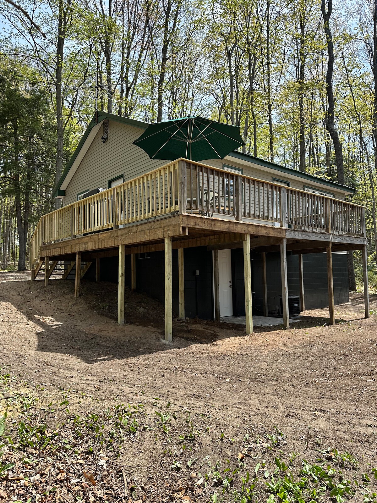 The cottage exterior is presented, featuring a wrap-around deck supported by wooden pillars. A large green umbrella provides shade. Surrounding trees contribute to a natural setting, and the ground beneath the deck is clear of foliage.