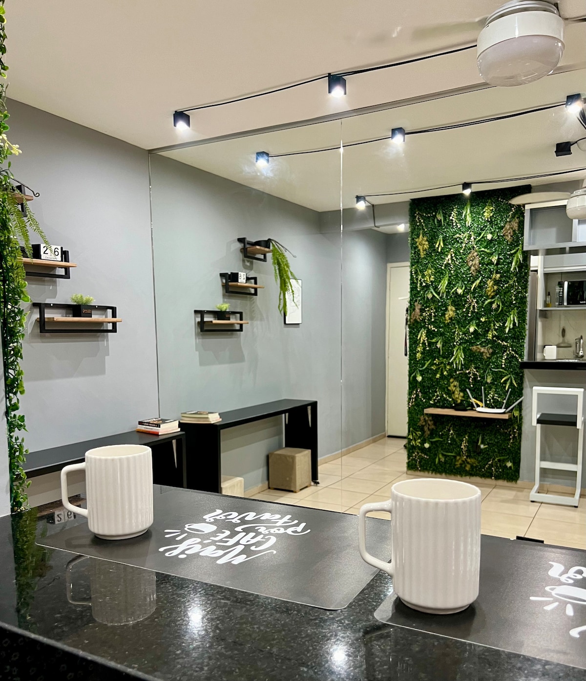 A modern kitchen area showcases a black countertop with two white mugs placed atop a textured mat. Green plant decor is displayed on wall-mounted shelves, while a vibrant green wall adds a refreshing touch to the space. A subtle lighting design enhances the overall ambiance.