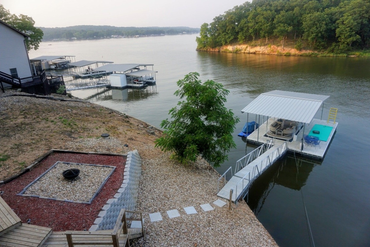The outdoor area features a gravel pathway leading to a new concrete dock with a swimming platform and a wet step ladder. A fire pit is surrounded by landscaping stones, and the serene lake view is framed by green trees on the opposite shore.