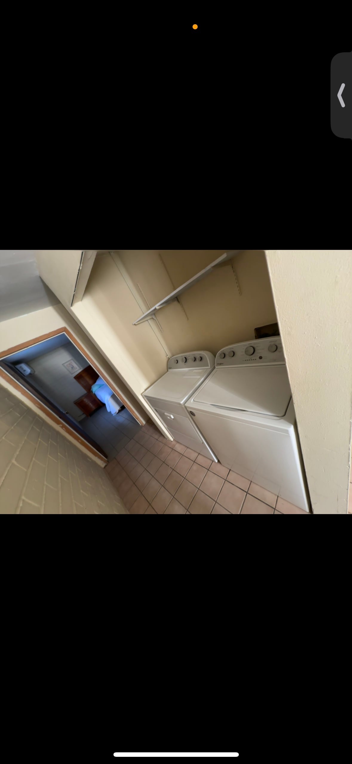 A functional laundry area is equipped with a washing machine and dryer, both positioned side by side. Above, a shelf provides additional storage space. The tiled floor complements the overall simplicity of the space, which leads into another room visible in the background.
