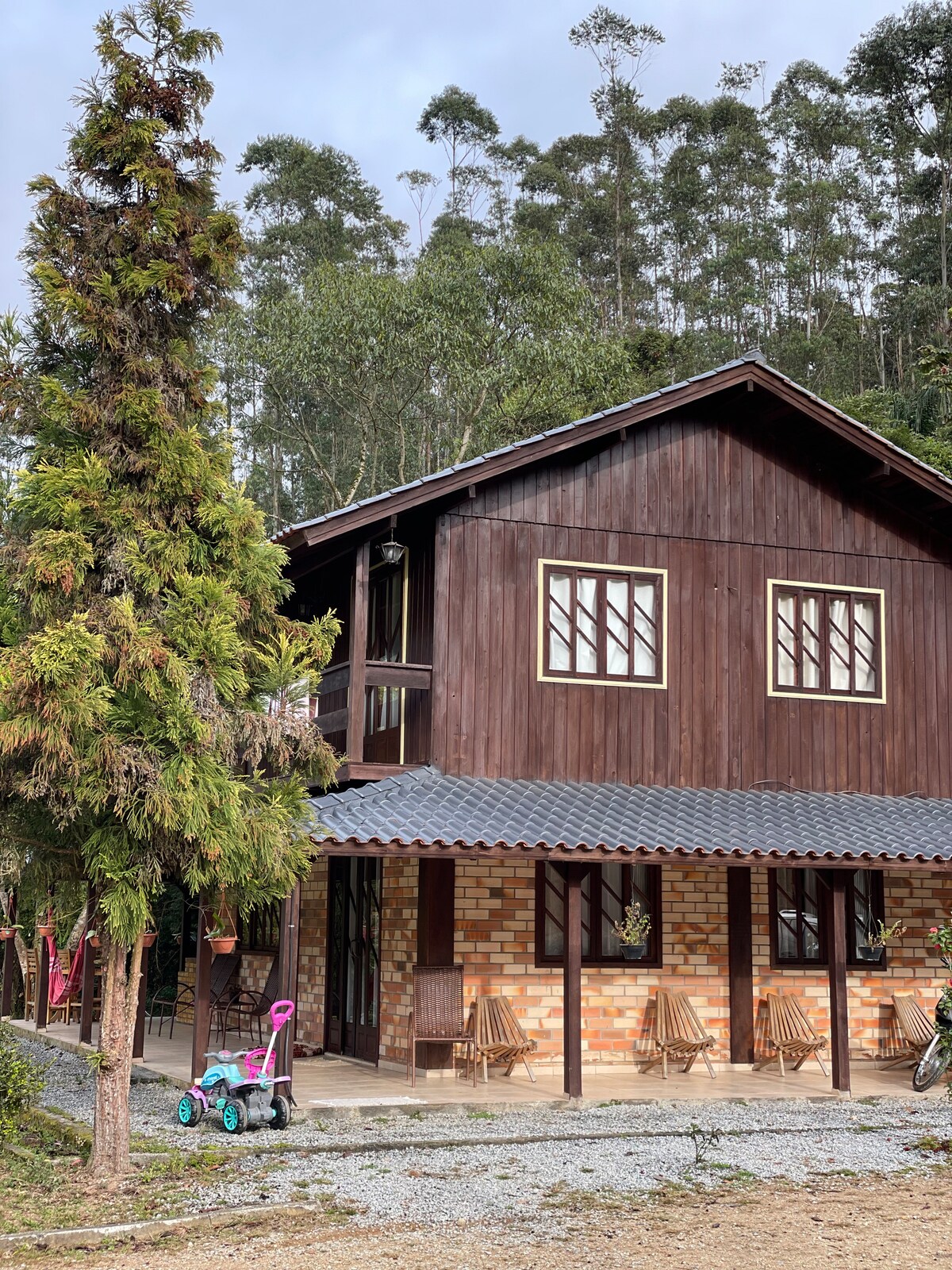 A two-story wooden house is surrounded by greenery, featuring a rustic exterior with large windows. A porch is equipped with wooden chairs, and a small children’s toy is placed in front. The gravel pathway leads to the entrance, enhancing the natural setting.