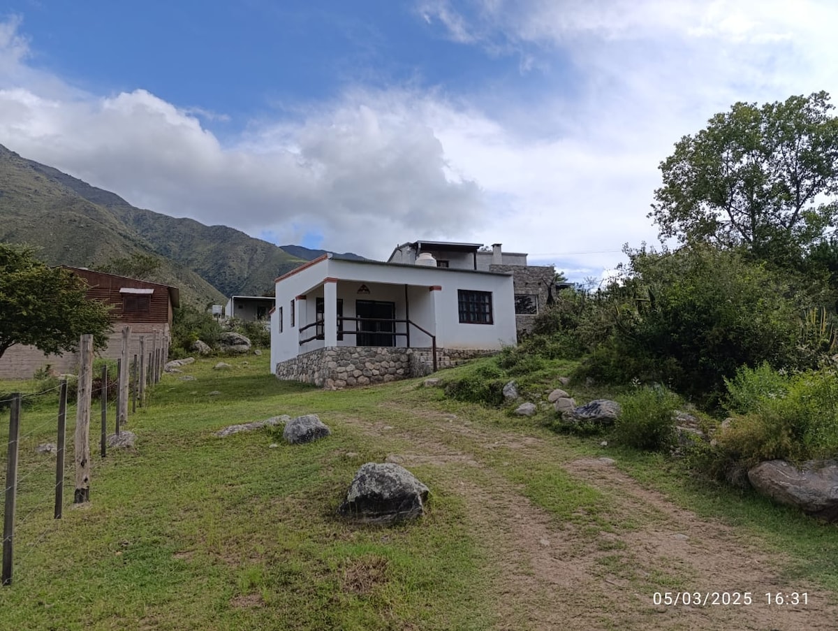 A single-story house is positioned on a gently sloping field, surrounded by greenery and boulders. The exterior features a stone foundation with white walls and a red roof. In the background, a mountain landscape rises under a partly cloudy sky.