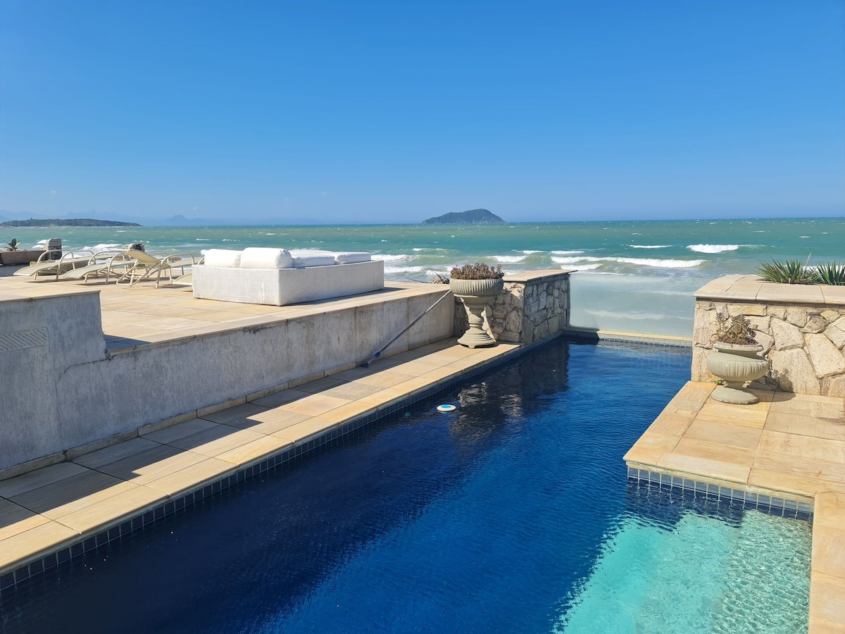 A serene pool area features a clear blue pool adjacent to a sandy beach. Lounge chairs are positioned on a spacious deck, with the ocean visible in the background. The tranquil water contrasts with the expansive sky, creating a peaceful seaside retreat.