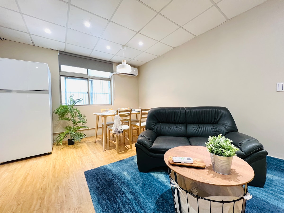 A well-lit living area is showcased, featuring a comfortable black sofa and a round wooden coffee table adorned with a small plant. A dining table with four chairs is visible in the background, alongside a large window that allows natural light to brighten the space.