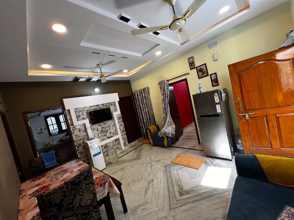 An open living area displays a dining table covered with a colorful tablecloth, surrounded by chairs. A kitchenette is visible, featuring a refrigerator and a ceiling fan. Light filters in through a window, and decorative curtains add a touch of character to the space.