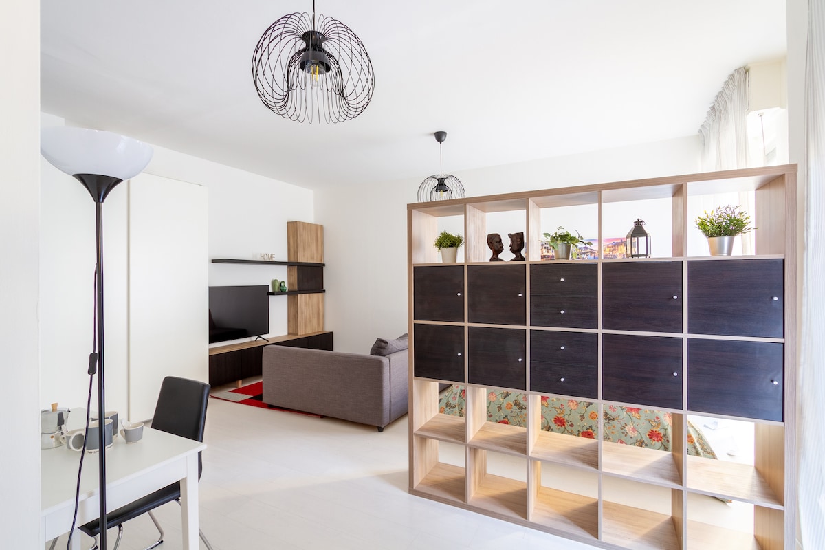 A modern living space is divided by a stylish bookshelf, featuring a mix of open and closed storage sections. A cozy sofa sits adjacent to a media unit, while a small dining table and lamp are positioned nearby. Natural light filters through the large windows.