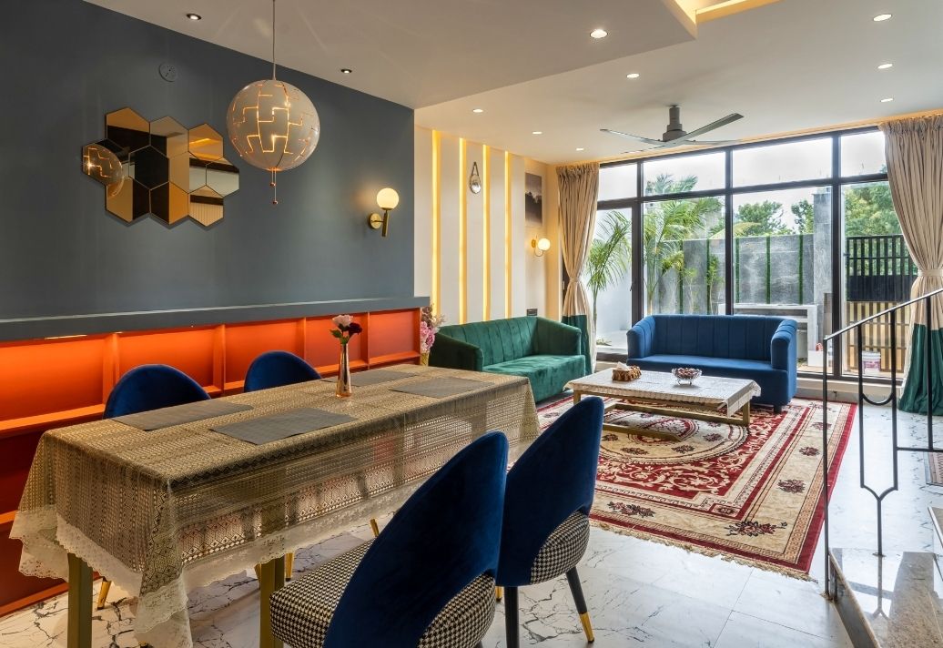 A modern living and dining area is showcased with a spacious dining table surrounded by blue chairs. A green and a light blue sofa are positioned nearby. Large windows allow natural light to illuminate the room, while decorative elements add warmth to the ambiance.