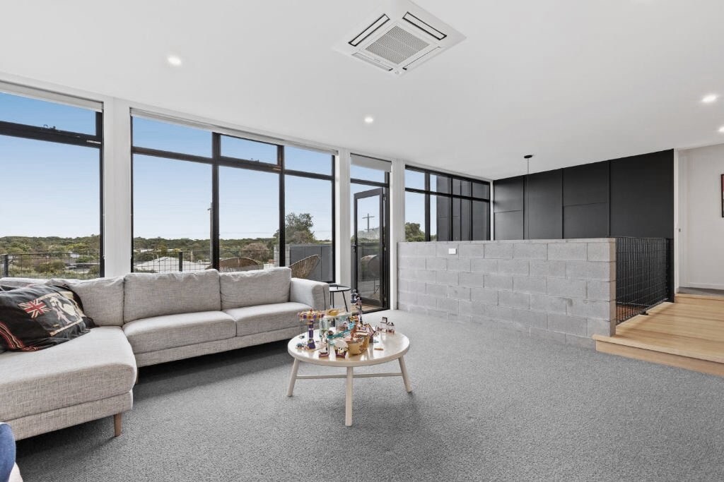 A light-filled living area features a spacious sectional sofa and a low coffee table adorned with decorative items. Large windows provide views of the surrounding landscape, while a minimalist design is complemented by a neutral carpet and warm wood accents.