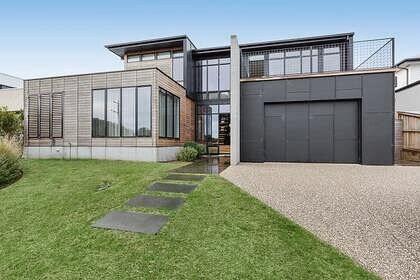 A modern architectural design is presented in this image of the exterior. A combination of materials, including wood and grey panels, characterizes the façade. Large windows are visible, allowing ample natural light, alongside a neatly landscaped front yard with a stone pathway leading to the entrance.