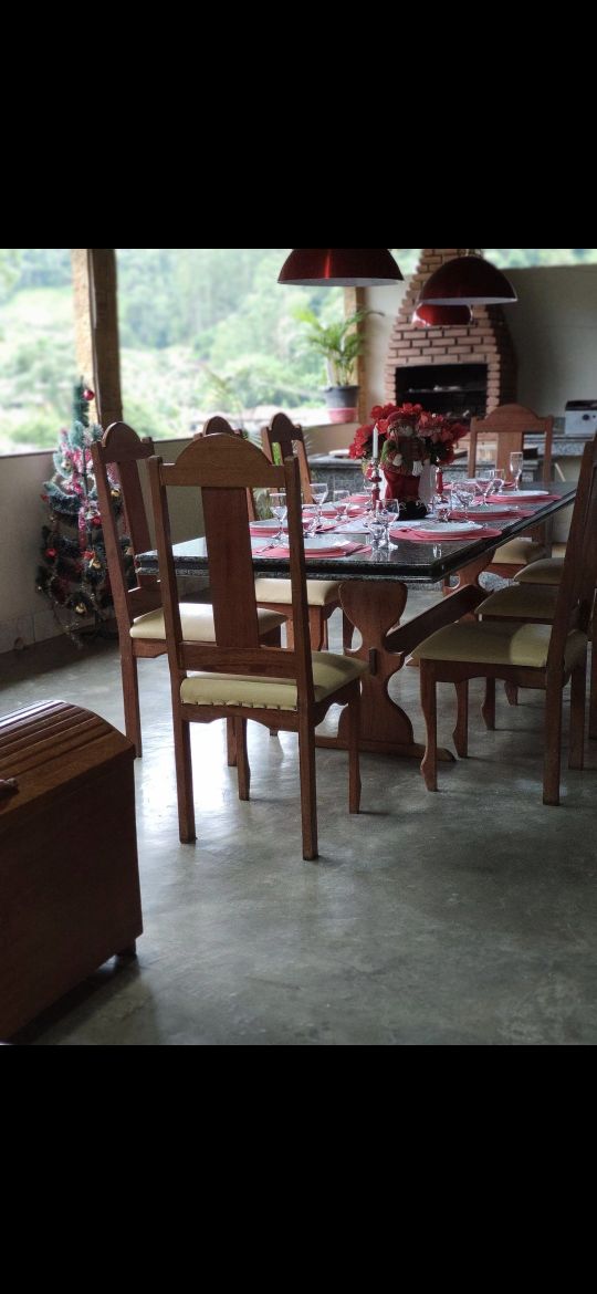 A dining area features a wooden table set for multiple guests, surrounded by sturdy chairs. Natural light enters through large windows, offering views of the surrounding greenery. A rustic brick barbecue is visible in the background, enhancing the space's functionality.