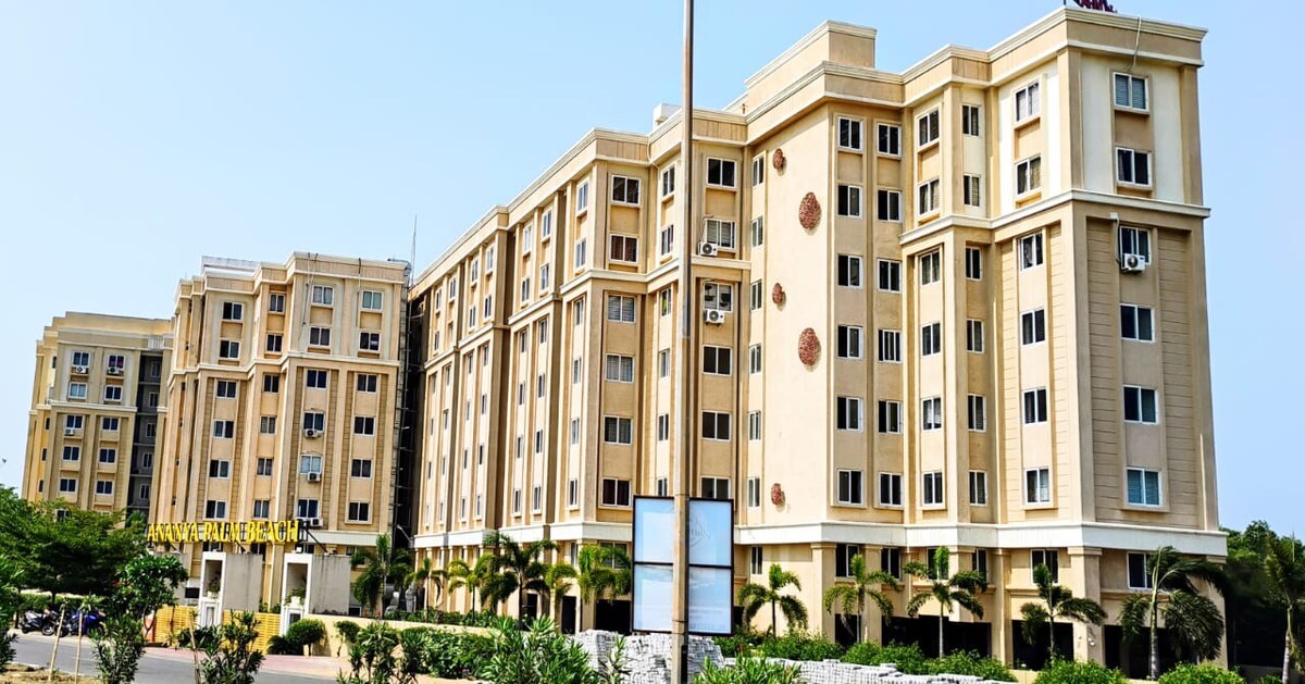 A modern multi-story building shows a beige exterior with numerous windows. Decorative elements adorn the facade, and palm trees are planted in the foreground. The structure is set against a clear blue sky, contributing to a welcoming environment.