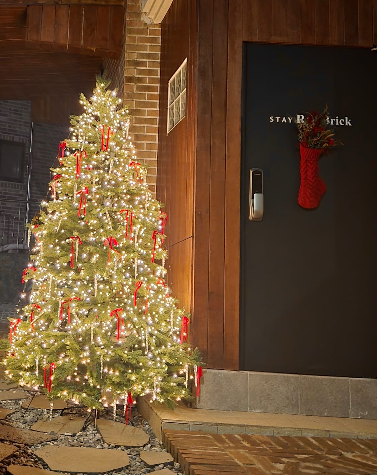 A festive outdoor area is displayed with a decorated Christmas tree adorned with lights and red ribbons, positioned next to an entrance. A holiday wreath hangs on the door, adding a seasonal touch to the warm wooden exterior.