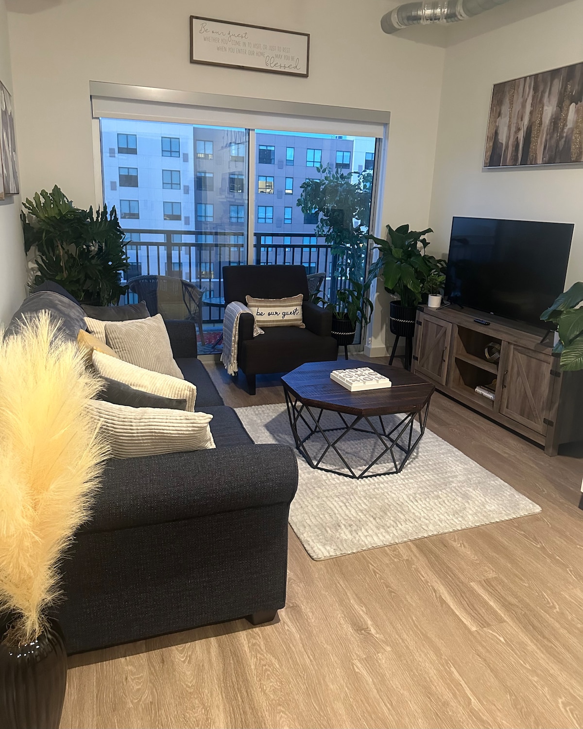 A cozy living area is featured, with a dark sectional sofa and an accent chair positioned around a round coffee table. Large windows allow natural light to brighten the space, while plants add a touch of greenery. A flat-screen TV is mounted on the wall for entertainment.