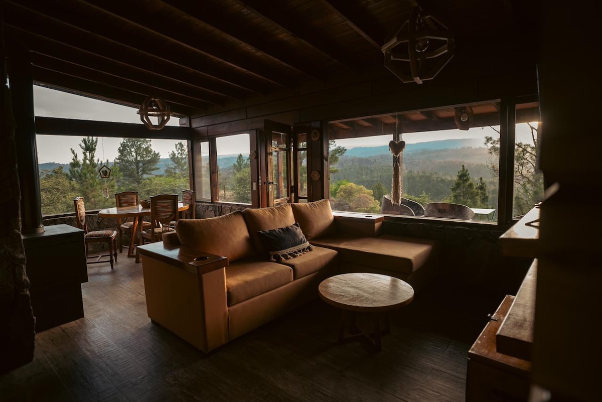 The living area features large windows that frame a breathtaking view of the surrounding nature. A spacious couch with neutral tones occupies one corner, accompanied by a wooden coffee table. A dining table for four is set nearby, enhancing the open, inviting space.