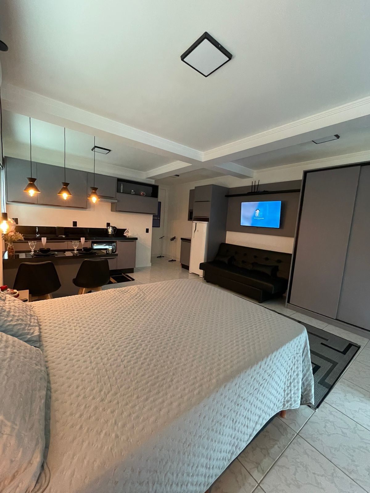 A modern studio apartment features a spacious bed with a neutral-colored quilt. The kitchenette is equipped with black chairs and soft lighting, while a television is mounted on the wall across from a sleek dark sofa. Light reflects off the tiled floor.