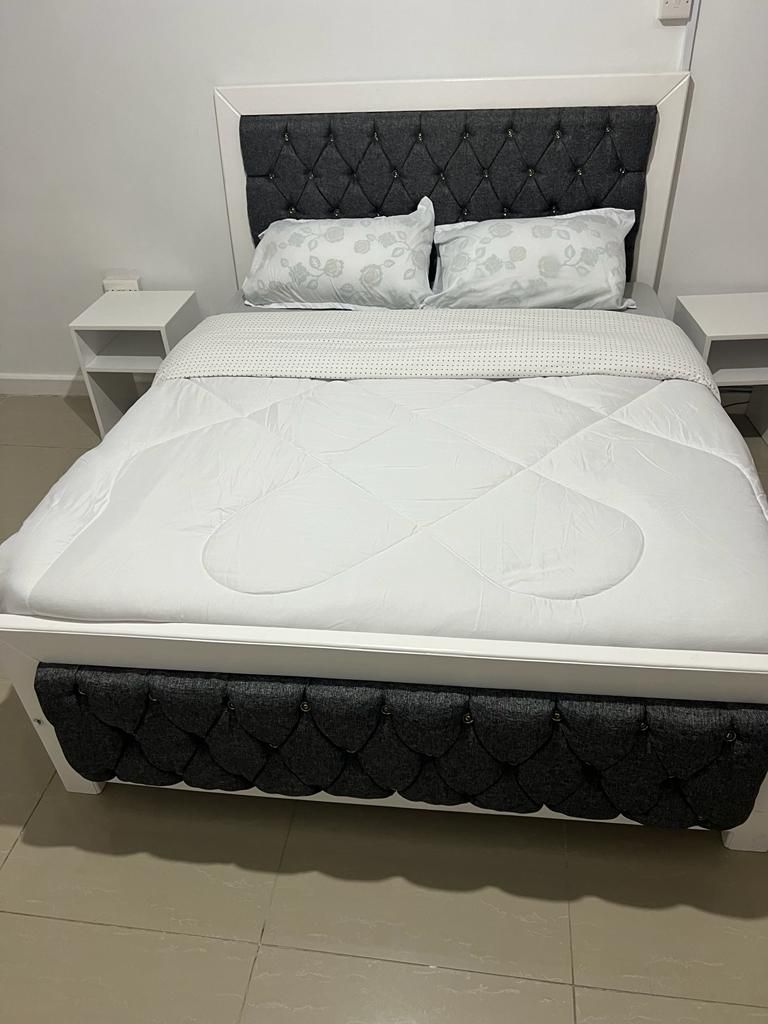 A spacious bed with a tufted headboard is positioned against a light wall, featuring two decorative pillows. Two nightstands are placed on either side of the bed, contributing to a clean and organized layout.