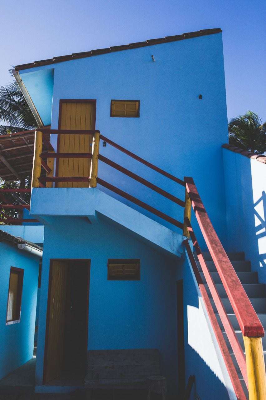 A bright blue exterior is highlighted by a staircase with a yellow railing leading to an upper level. Windows are set into the building, providing natural light, while a nearby palm tree adds a touch of greenery to the scene.