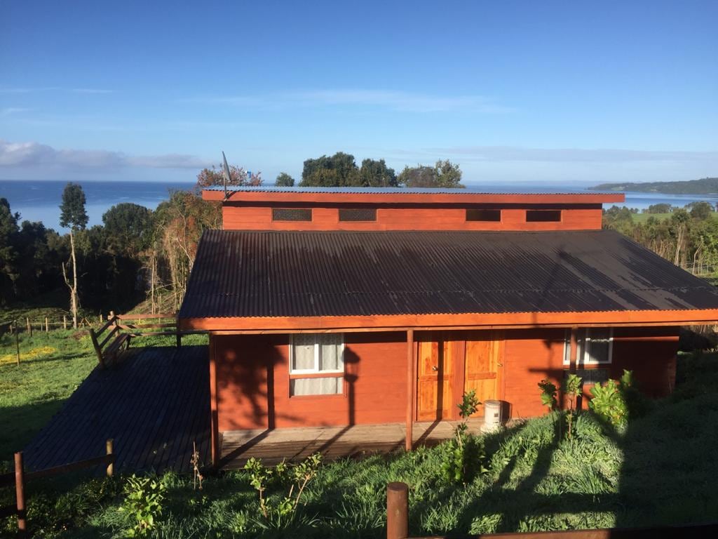 A side view of a rustic wooden cabin is presented, with large windows allowing natural light to enter. The cabin features a sloped roof, and a deck extends out towards a lush green landscape. In the background, the calm waters of a lake are visible under a clear sky.