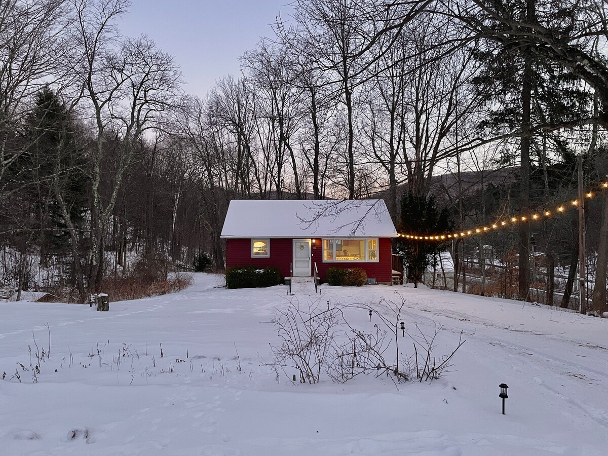 A charming red cottage is nestled among bare trees in a snow-covered landscape. Soft lighting from string lights glows above a clear path leading to the front door, which is framed by large windows. The tranquil scene evokes a serene winter ambiance.