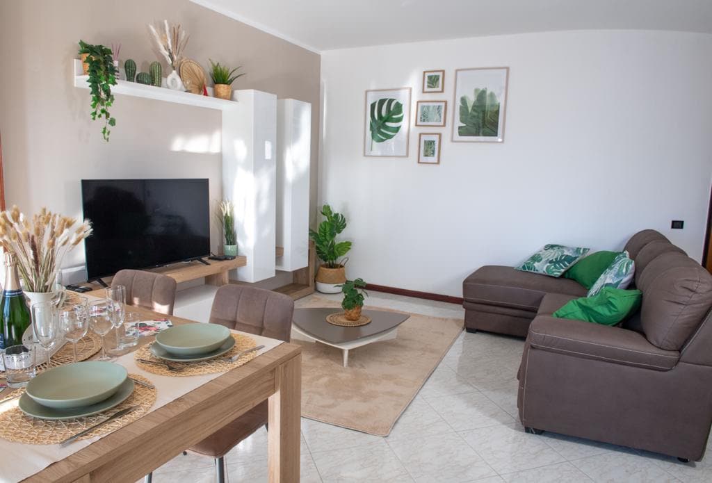 The living area features a cozy brown sectional sofa adorned with green accent pillows. A flat-screen TV rests on a white console while framed botanical prints decorate the wall. A dining table, set with plates and glassware, complements the inviting space, filled with natural light.