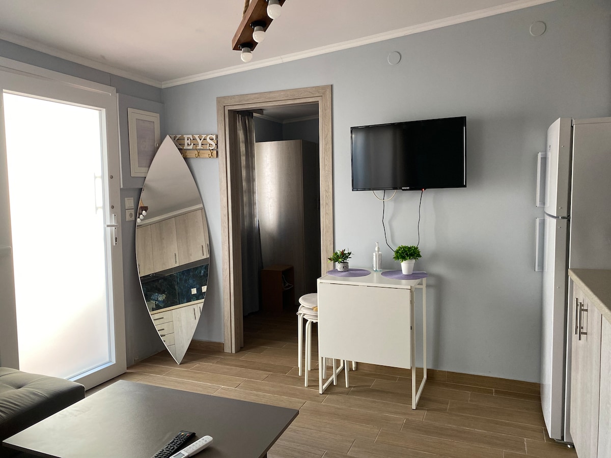 A modern kitchen area is visible, featuring a sleek dining table with two chairs. A large mirror reflects light, while a wall-mounted television adds convenience. Light wooden flooring complements the neutral wall tones, enhancing the overall spacious feel of the area.