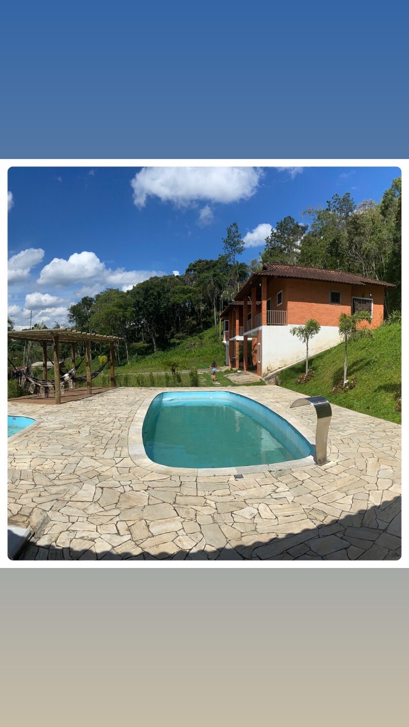 A clear view of a rectangular pool is presented, surrounded by a stone patio. Lush greenery and trees create a serene backdrop, while a two-story building is visible in the distance, featuring warm brick tones. Fluffy clouds dot a bright blue sky.