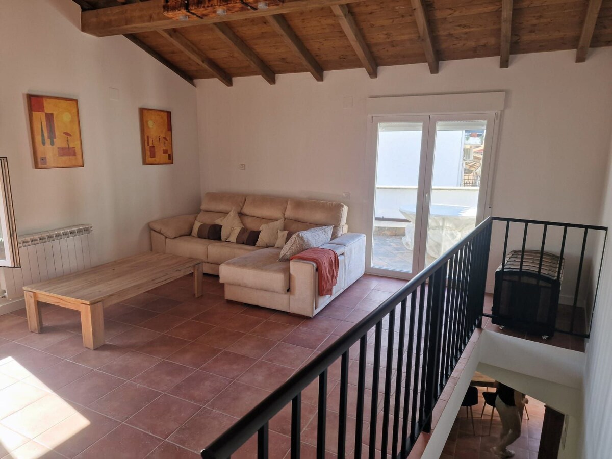 The living area features a comfortable sectional sofa arranged around a wooden coffee table. A staircase with a black railing is visible, leading to a lower level. Natural light fills the space through large windows, highlighting the wooden ceiling beams and tiled floor.