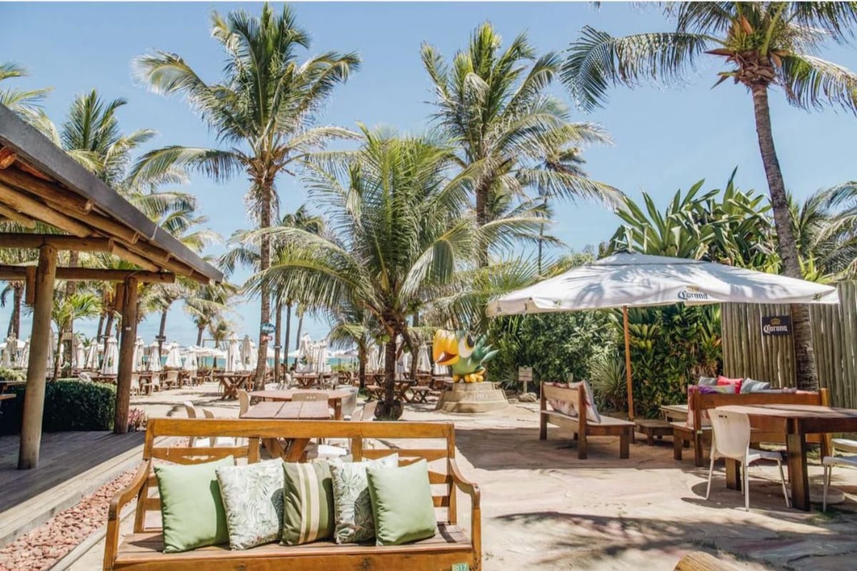 A spacious outdoor area is surrounded by lush palm trees and tropical plants. Wooden benches are arranged for seating, with white umbrellas providing shade. Sun loungers are visible in the background, creating a relaxed atmosphere near the beach.