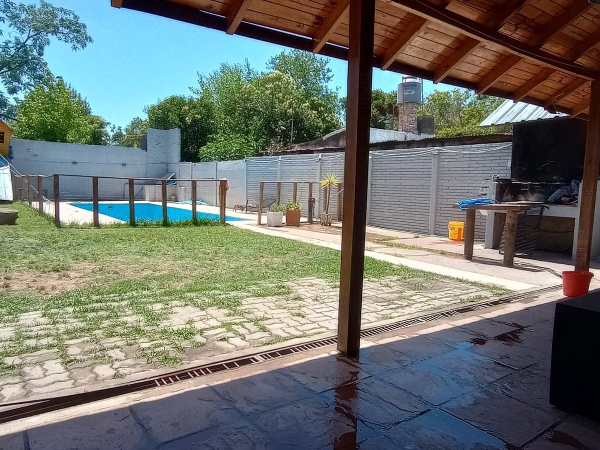 An outdoor view is presented, featuring a swimming pool surrounded by a wooden deck. Lush greenery and trees are visible in the background, with a barbecue area positioned nearby. The setting is designed for relaxation, with an open structure providing a connection to the landscape.