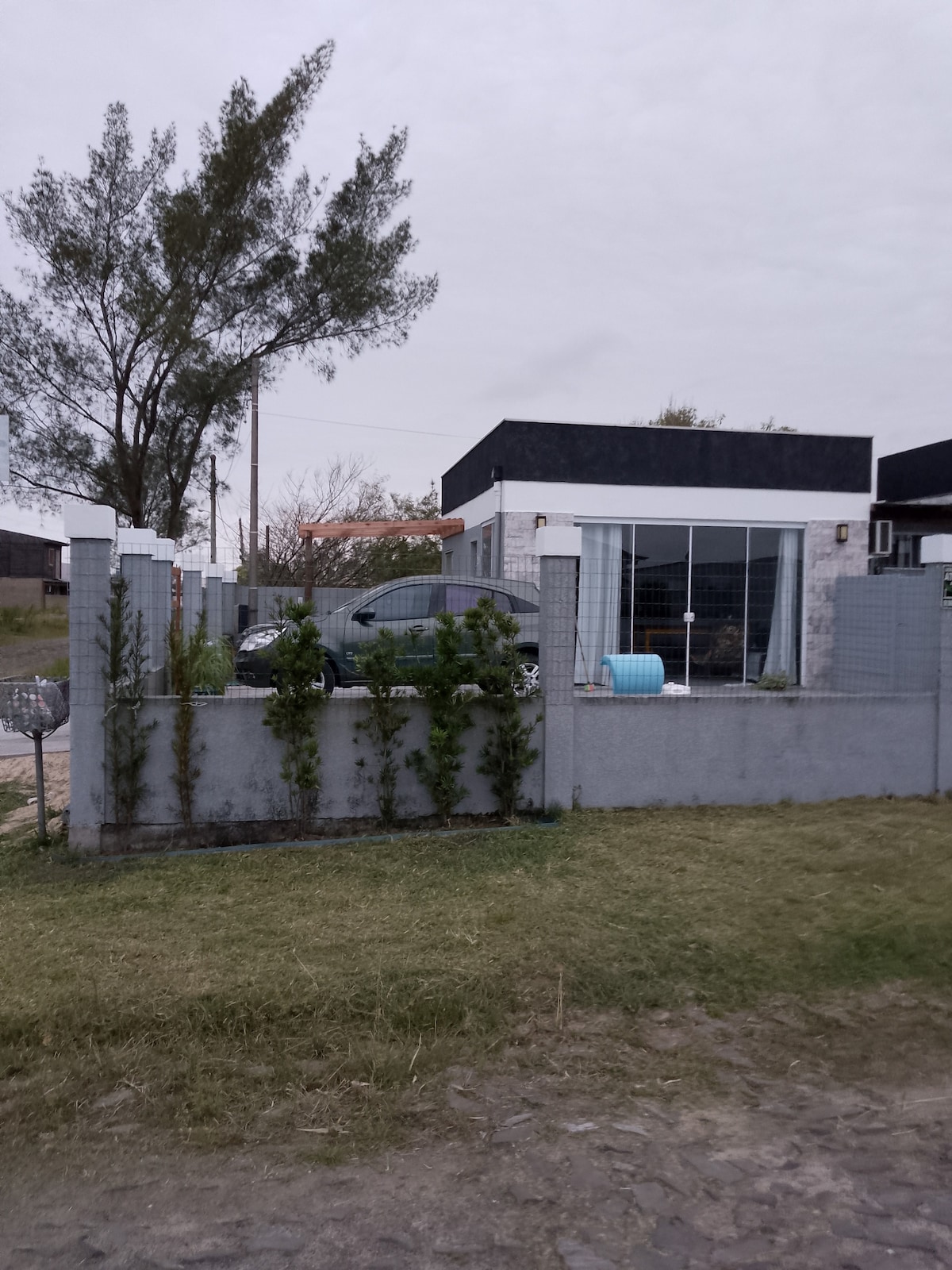 A modern house is situated on a corner lot, featuring a secure fence and landscaped garden. A car is parked in the driveway, and large glass windows allow glimpses of the interior space. Surrounding greenery adds to the outdoor appeal.