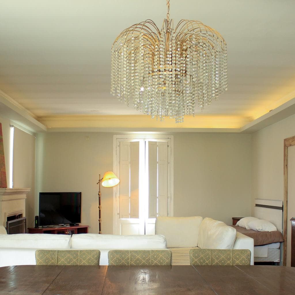 A spacious living area features a large dining table with ornate chairs, complemented by a modern chandelier overhead. A comfortable sofa is positioned near the entertainment unit, while natural light enters through double windows with classic shutters. Neutral walls enhance the inviting ambiance.