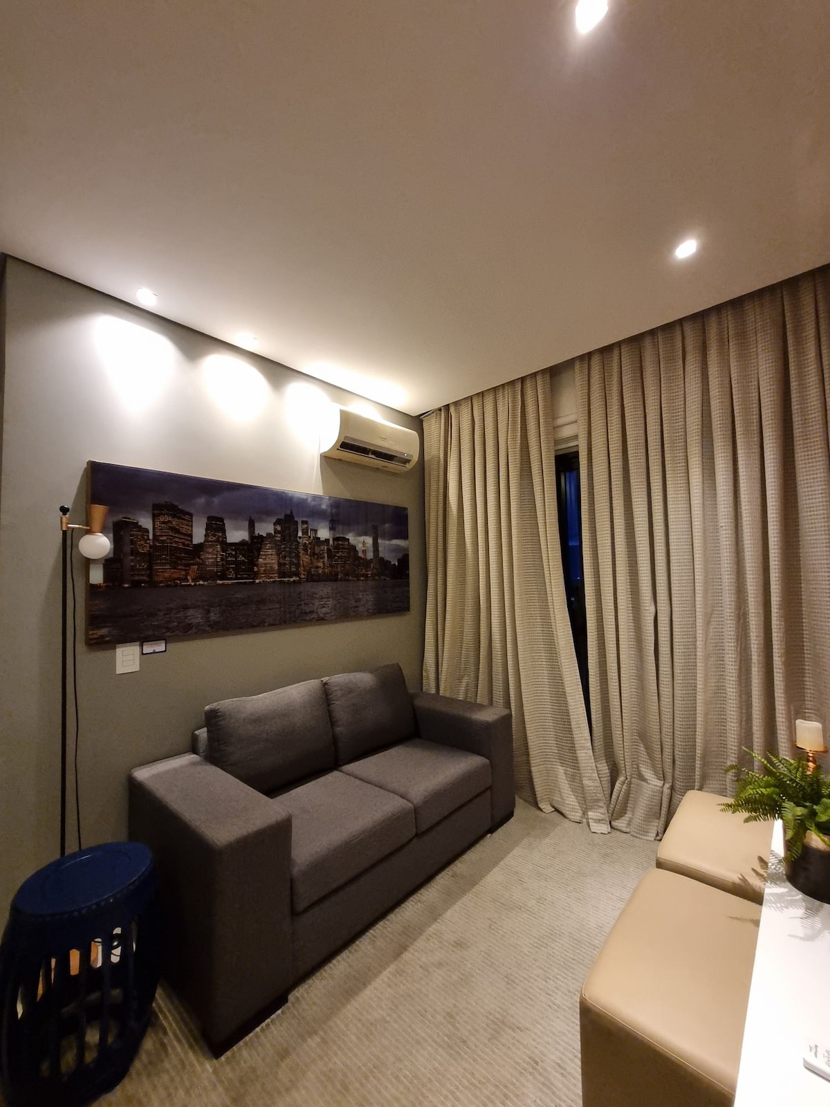 The living area features a modern gray sofa against a wall adorned with a cityscape artwork. Soft lighting is provided by recessed ceiling lights, and floor-to-ceiling curtains frame the window. A small, round table with a potted plant is positioned nearby.