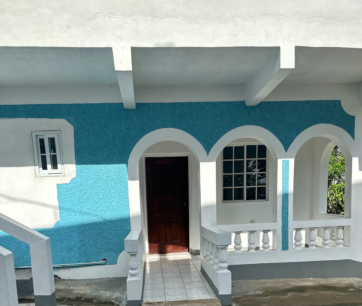 The exterior features a bright blue wall with white accents and arches, providing a clean and inviting entrance. A wooden door leads into the cottage, with several windows allowing natural light to enter. A stairway is visible leading to the upper level.