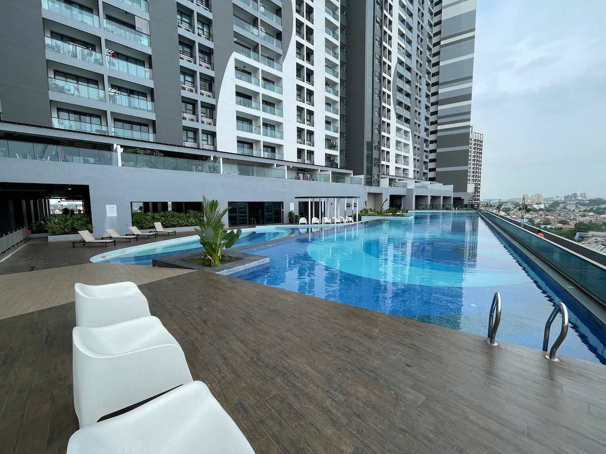 An inviting swimming pool area features a cascading infinity edge, surrounded by loungers for relaxation. The modern building's glass facade reflects the sky, while lush greenery enhances the tranquil atmosphere. Poolside access leads to a spacious deck overlooking the city.