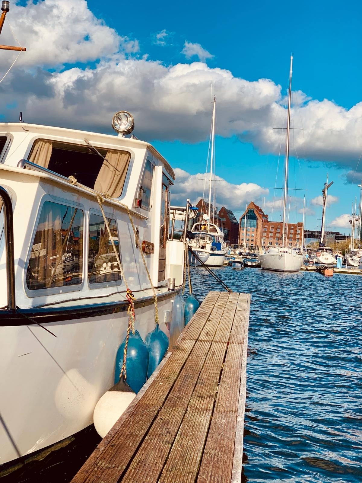A charming boat is docked alongside a wooden pier, with blue fenders hanging from its side. The water reflects the sky and nearby sailboats, while fluffy white clouds drift overhead, creating a tranquil harbor scene.