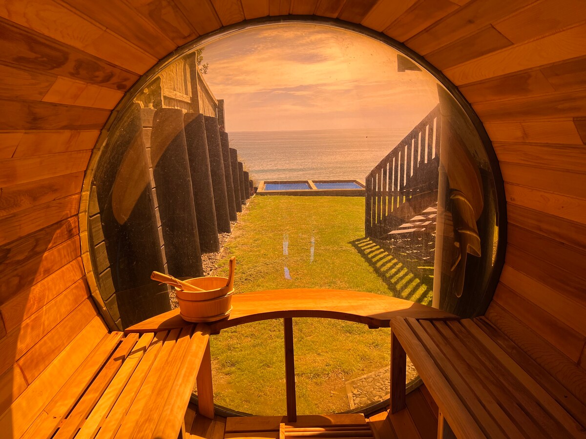 A view of the outdoor landscape is captured through a curved window of a wooden sauna, showcasing a grassy area leading to steps down toward the beach. Soft sunlight bathes the scene, with the ocean visible in the background.