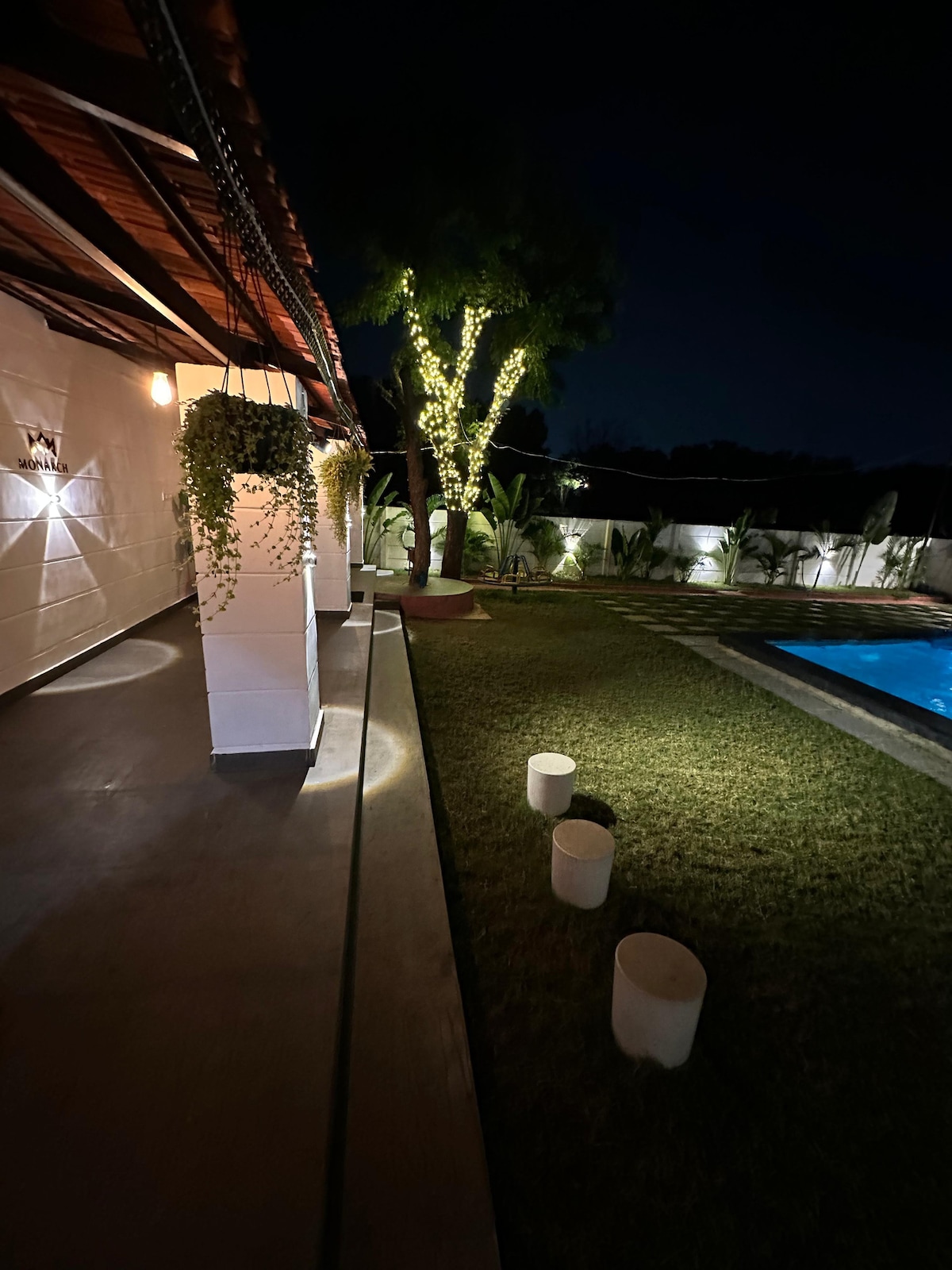 A nighttime view of the property highlights the illuminated pathway leading to the pool area. Soft lighting is provided by strategically placed fixtures along the walls and around a nearby tree, enhancing the lush greenery of the manicured lawn.