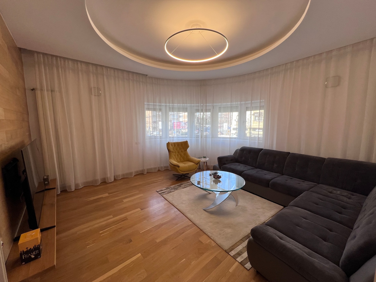 A spacious living area features a large sectional sofa and a distinctive yellow chair. Soft curtains frame the windows, allowing natural light to fill the room. A circular ceiling light offers subtle illumination. A round glass coffee table rests on a light area rug.