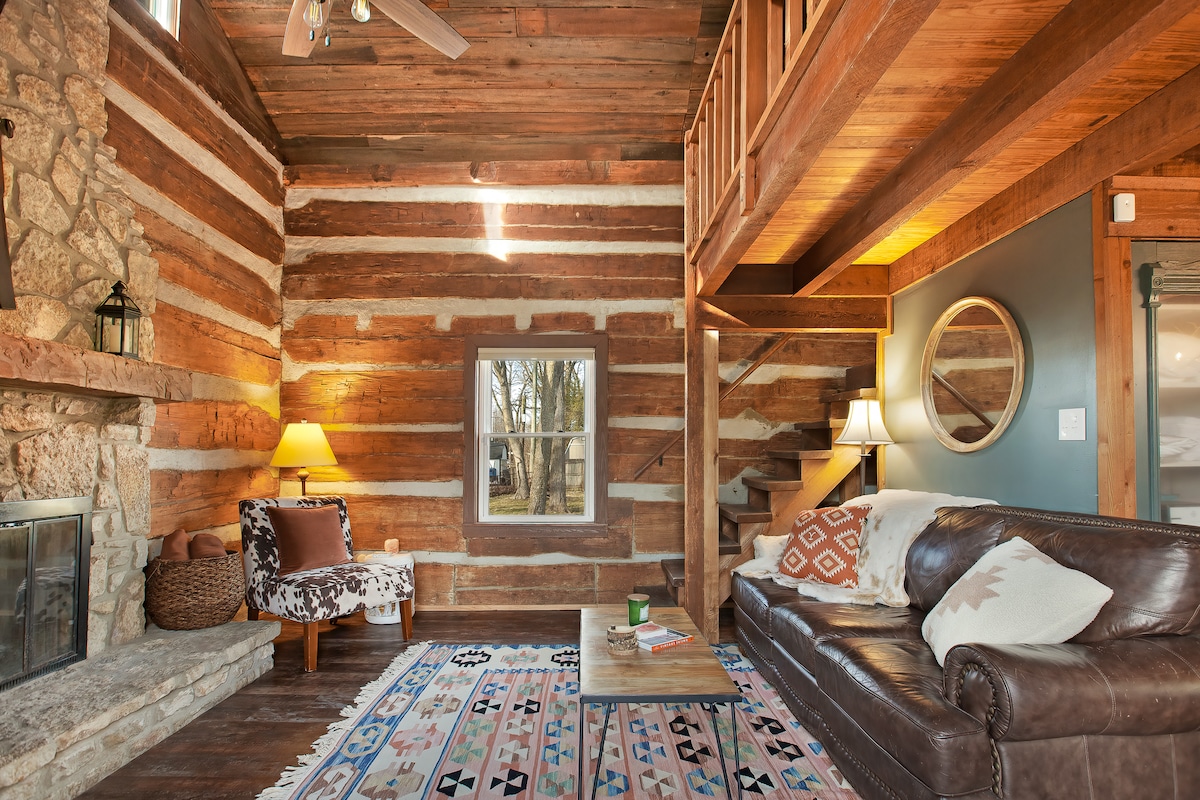 The living area features exposed wooden beams and a large stone fireplace, complemented by a cozy leather sofa and a patterned area rug. A staircase leads to the loft bedroom, while abundant natural light filters through the nearby window, enhancing the rustic charm.
