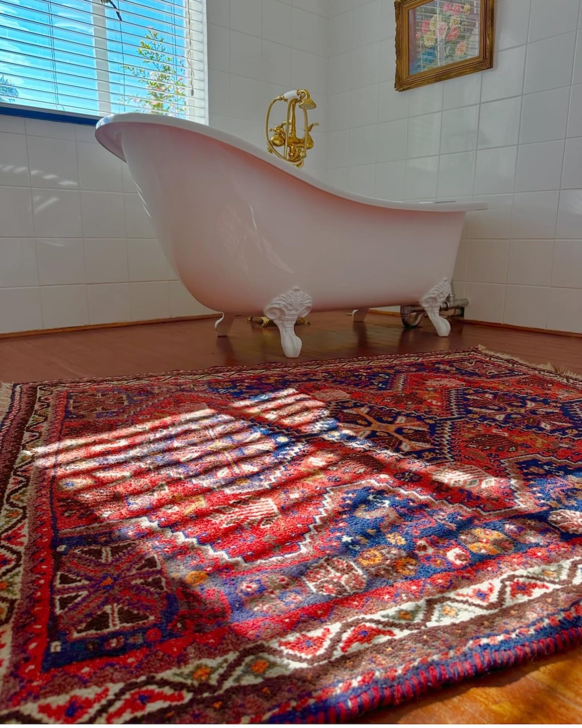 A freestanding bathtub with ornate golden fixtures is positioned against a tiled wall, allowing natural light to illuminate the space. A richly patterned rug in hues of red, blue, and cream provides warmth and texture on the wooden floor.