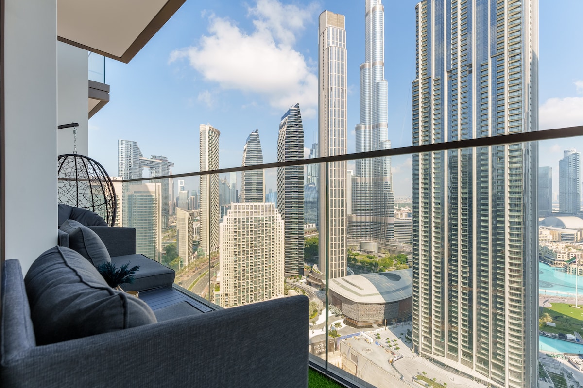 A modern balcony features a comfortable grey sofa, providing a perfect vantage point of the city skyline. Towering skyscrapers, including the Burj Khalifa, dominate the landscape under a clear blue sky, offering a striking contrast to the urban scene below.