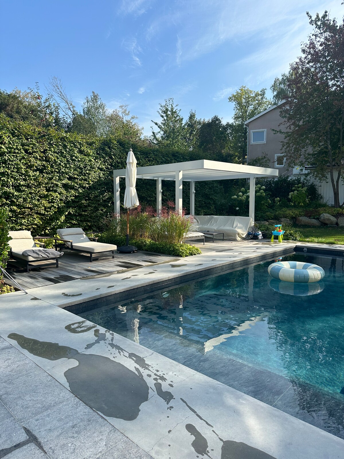 A spacious outdoor pool area features a crystal-clear water surface, surrounded by lush greenery. Two lounge chairs are positioned on a wooden deck beside the pool, offering a shaded pergola. A floating ring and vibrant flowers add color, enhancing the inviting atmosphere.