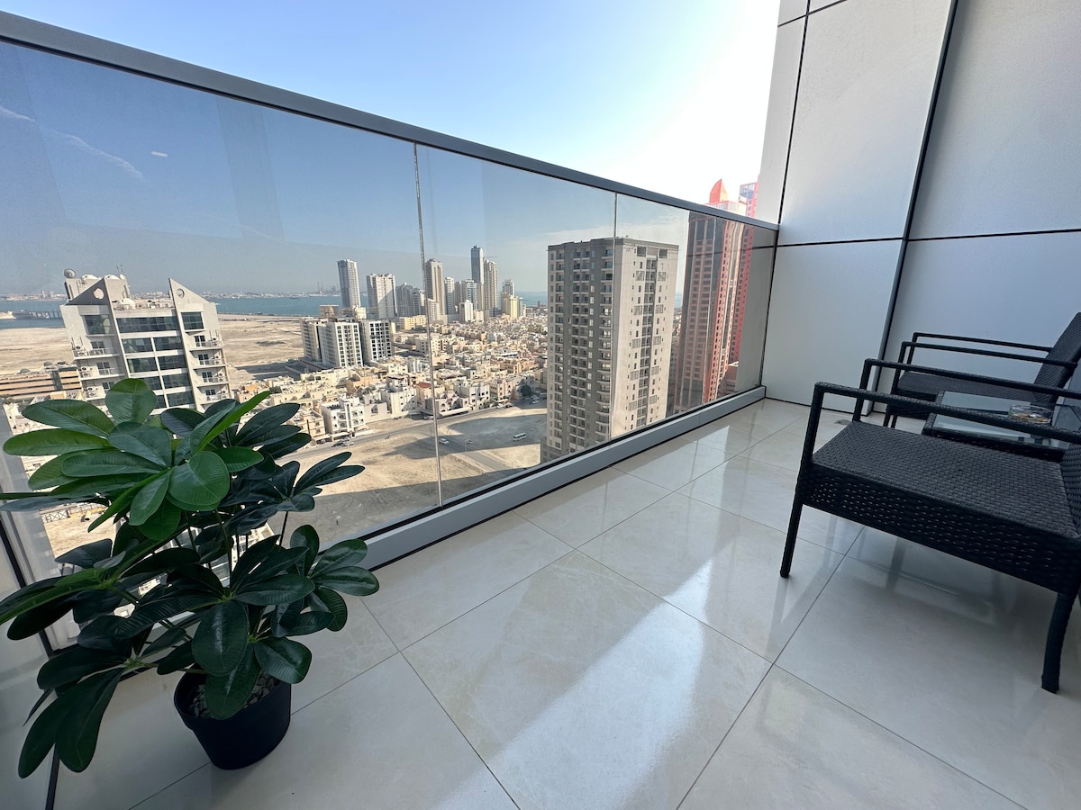 A private balcony is visible, featuring two comfortable chairs and a potted plant. The glass railing offers an unobstructed view of the surrounding cityscape and coastline, allowing ample natural light to fill the space.