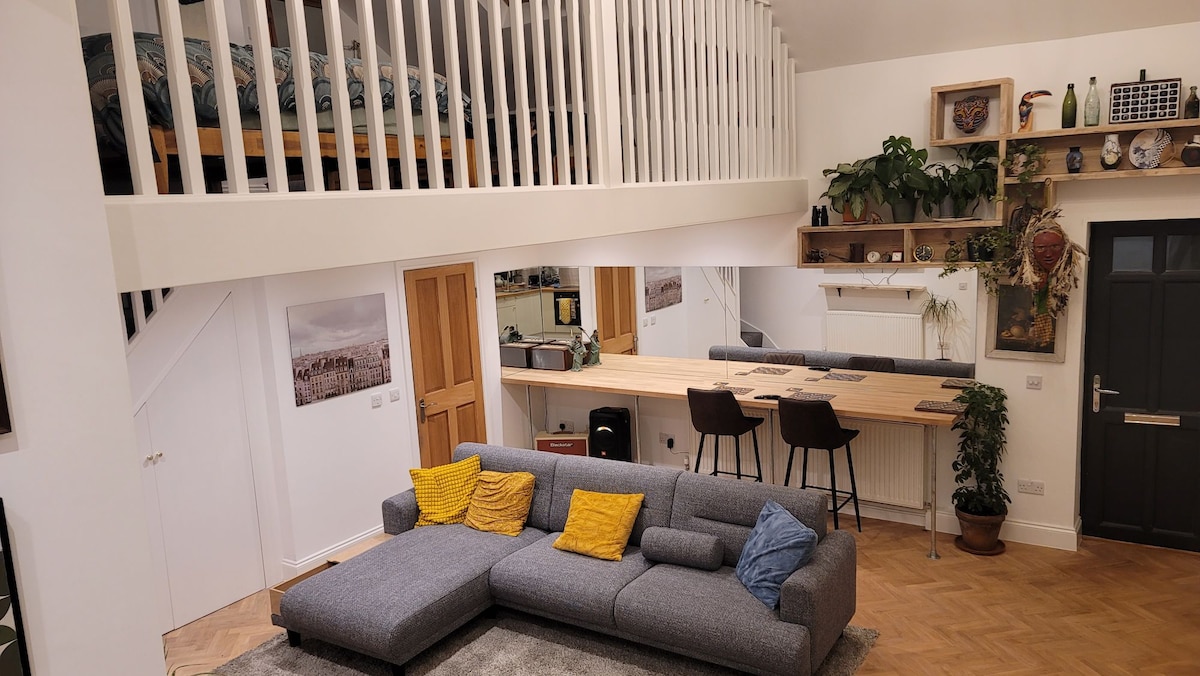 An open living space features a spacious gray sectional sofa adorned with yellow cushions. A wooden bar counter accommodates two stools, while plants decorate the shelves above. A lofted sleeping area is visible, with a cozy bed partially shown. Warm light fills the room.