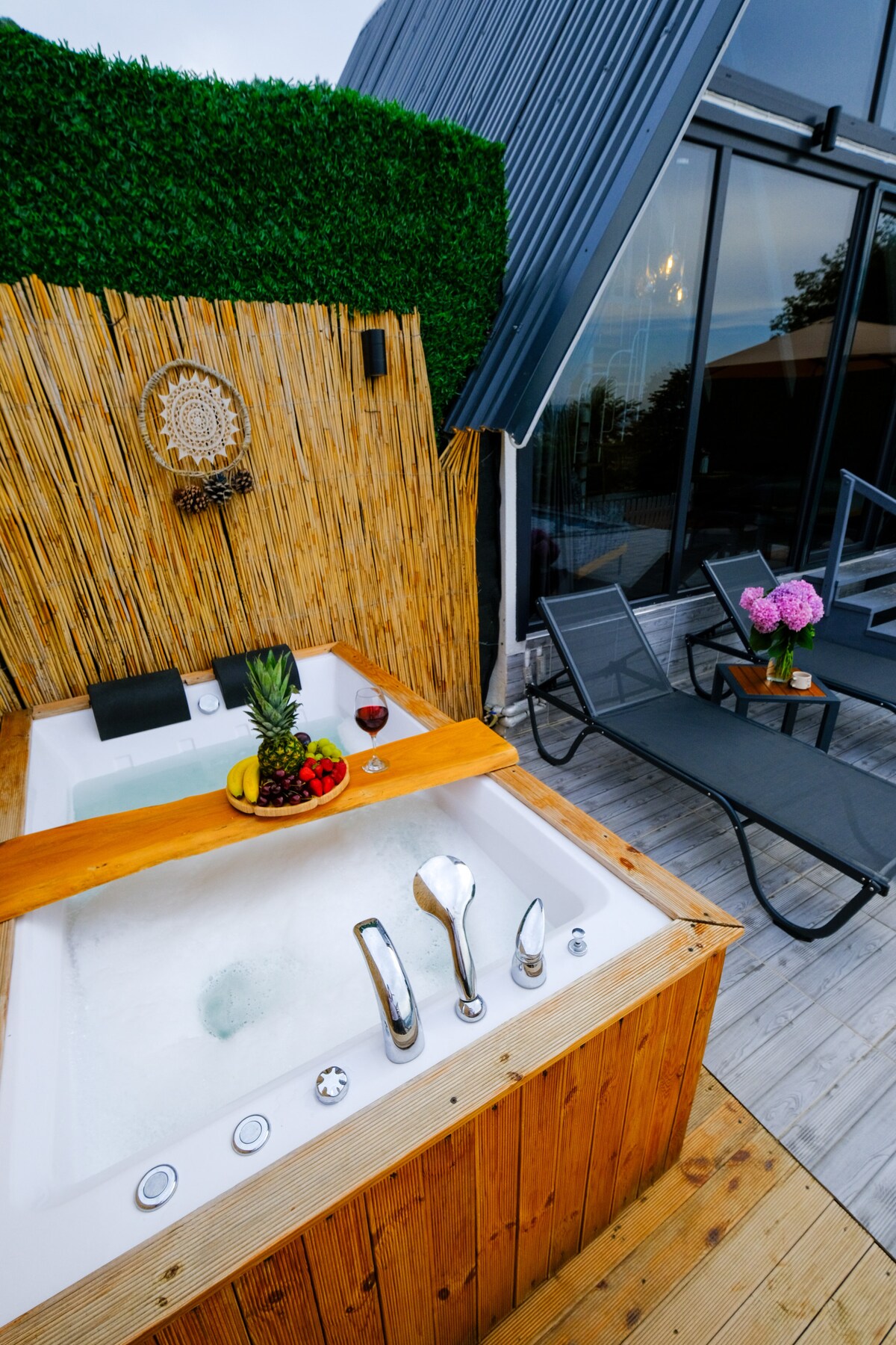 An outdoor jetted tub is surrounded by wooden paneling and lush greenery, offering a private space for relaxation. A wooden tray holds fresh fruit and a glass of red wine. In the background, lounge chairs provide additional seating options near the tub.