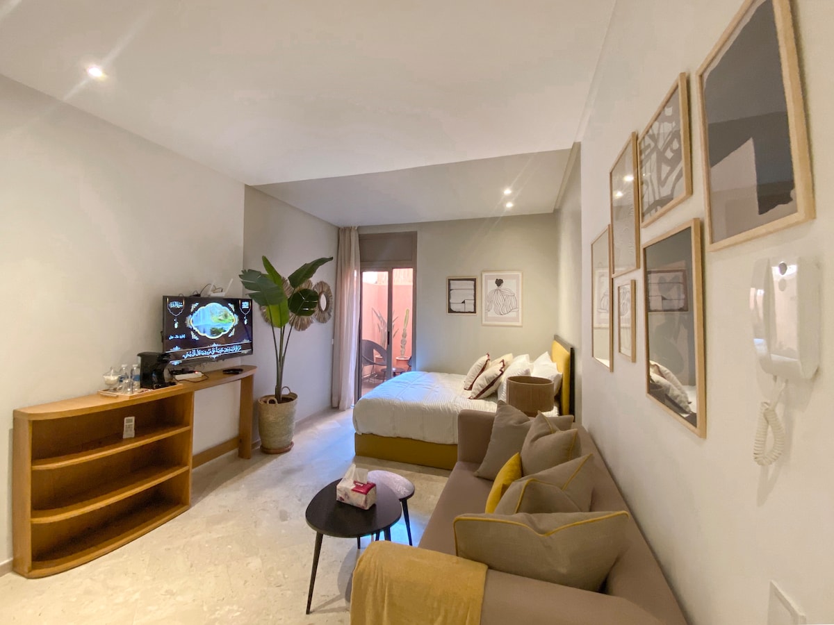 A modern living space features a comfortable corner sofa and a queen bed with light bedding. A TV is positioned on a wooden shelf, while framed art hangs on the neutral walls. Natural light enters through large windows, enhancing the welcoming atmosphere.