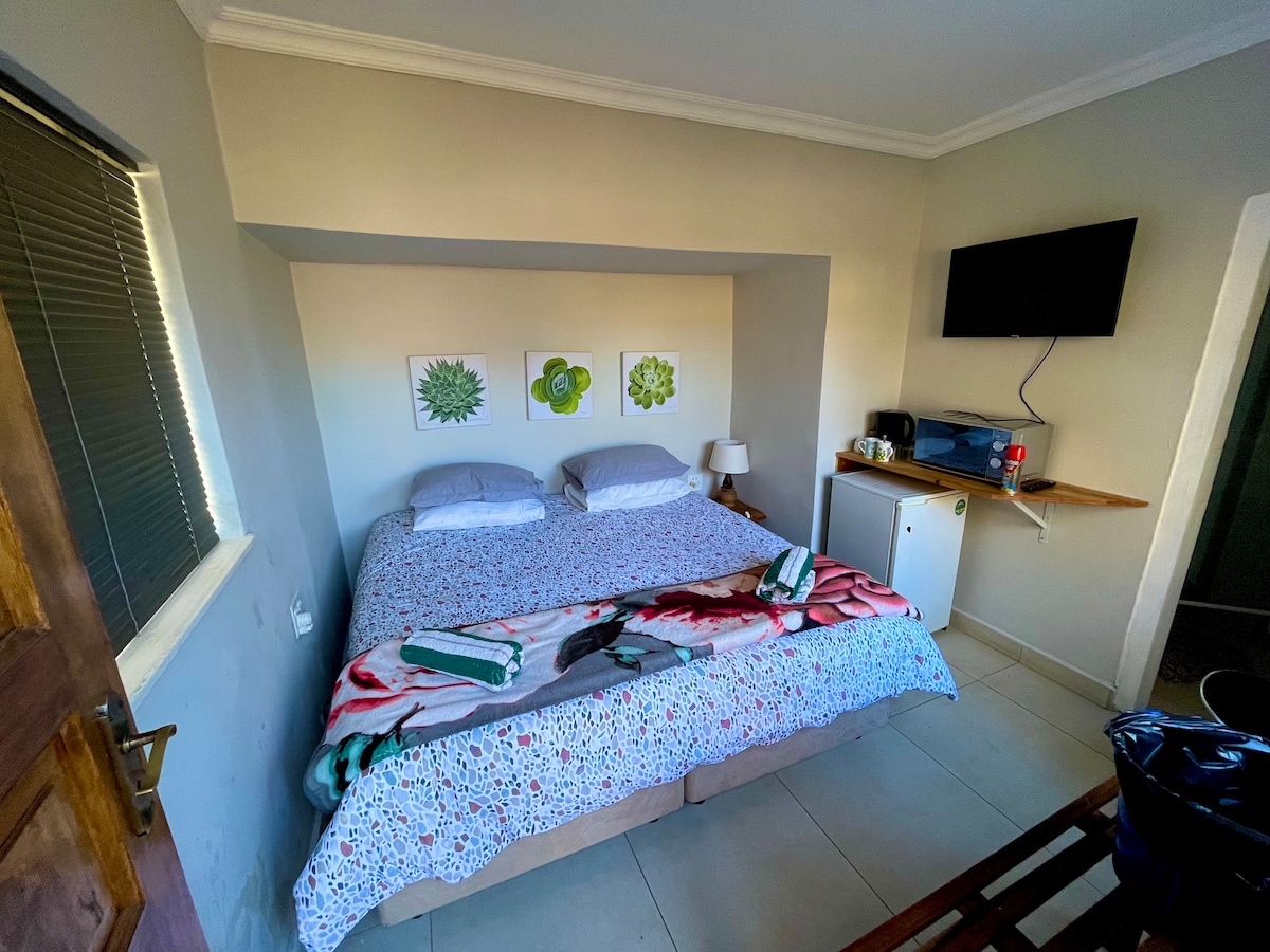 A king-bed arrangement is situated in a light-filled room, with artwork featuring botanical prints displayed above the bed. A small nightstand holds a lamp, and a television is mounted on the wall. A small refrigerator and coffee-making station are positioned on a shelf nearby.