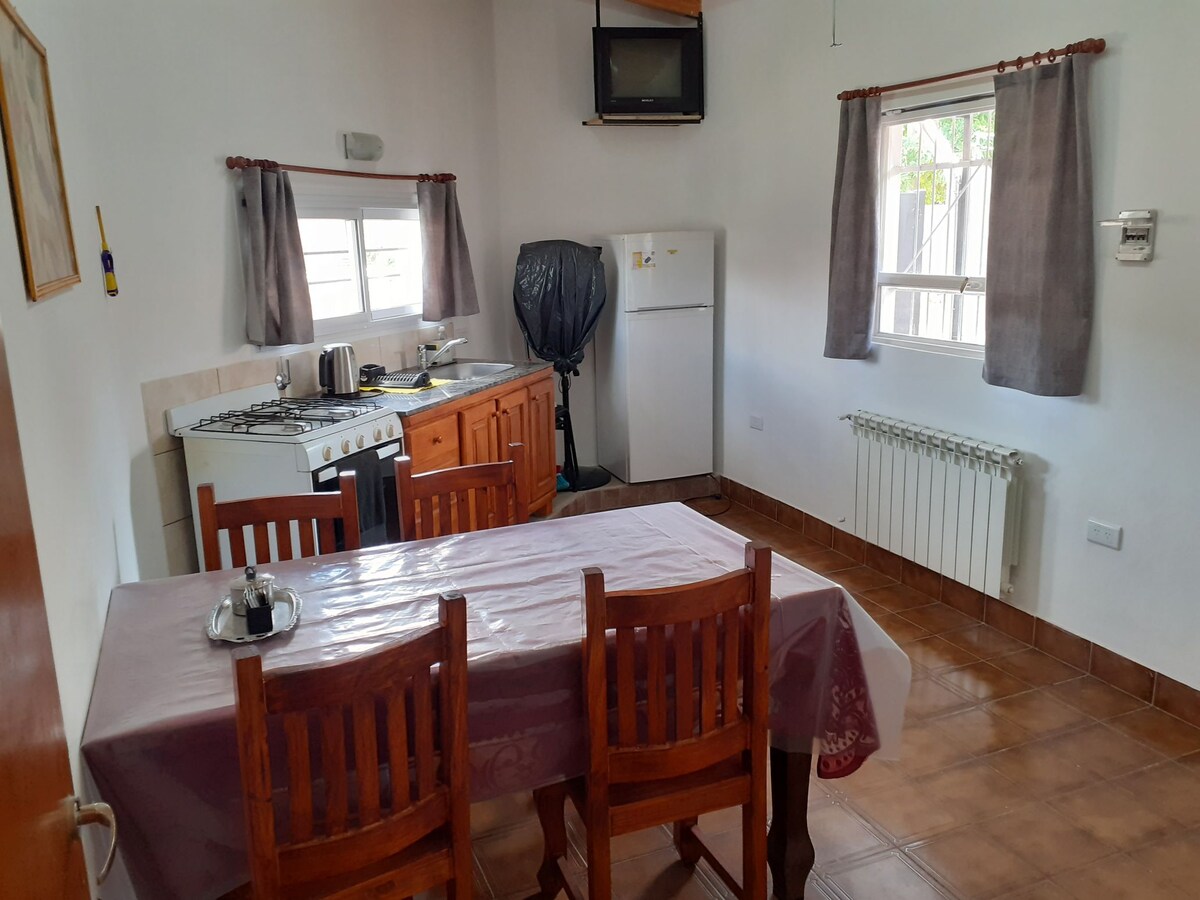 A dining area features a wooden table surrounded by five chairs, illuminated by natural light from nearby windows. A well-equipped kitchen is visible, showcasing a gas stove, refrigerator, and essentials. Neutral walls and tiled flooring contribute to a clean and functional space.
